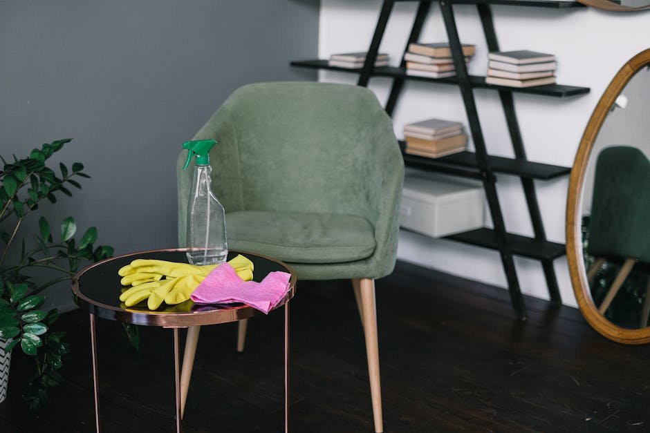 A room with a green upholstered armchair positioned next to a small round black table with a copper-colored rim, which holds yellow rubber cleaning gloves, a pink cleaning cloth, and a clear spray bot