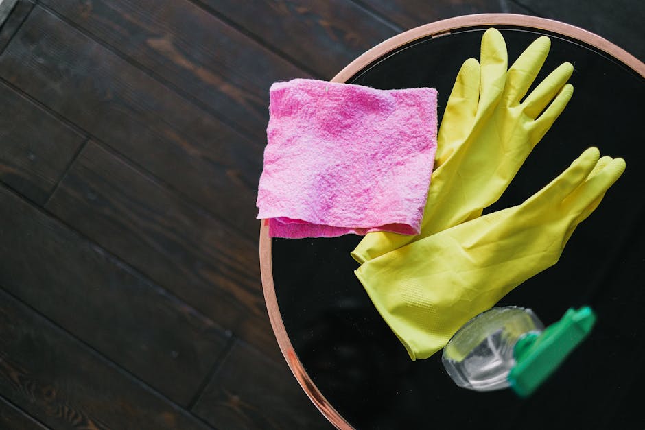 A room with a green upholstered armchair positioned next to a small round black table with a copper-colored rim, which holds yellow rubber cleaning gloves, a pink cleaning cloth, and a clear spray bot