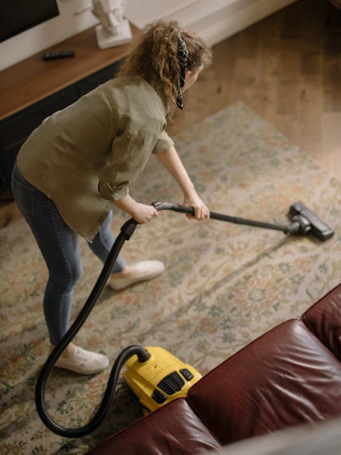 A child wearing beige trousers, white sneakers, and a beige jacket is vacuuming a patterned area rug with a yellow vacuum cleaner. The vacuum cleaner has a black hose and a slim metal wand, and it is 